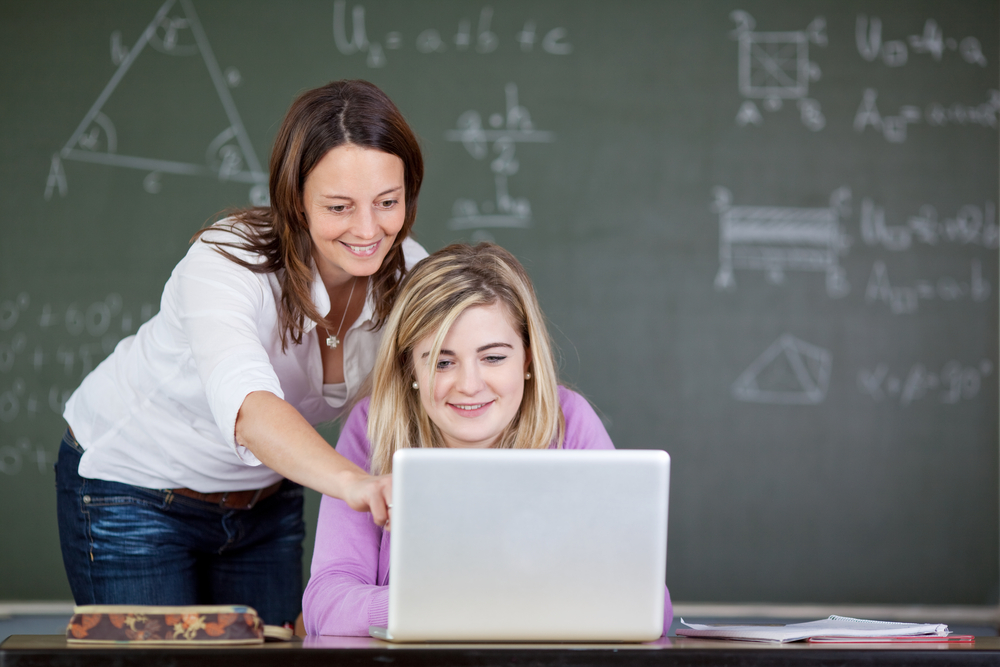 Female teacher assisting student in using laptop at desk in classroom.